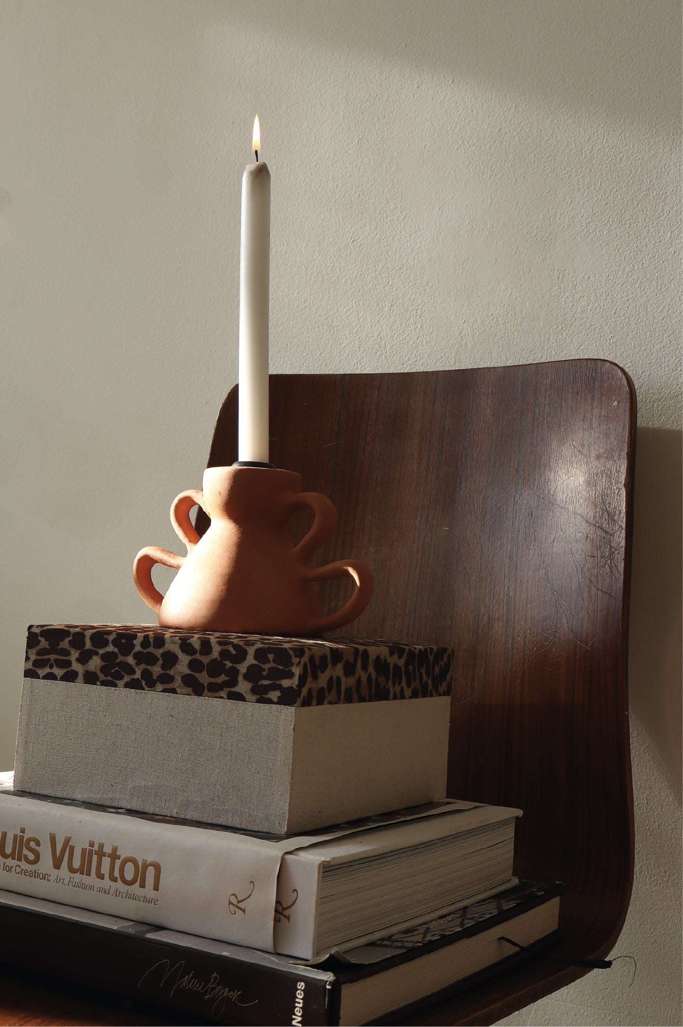 Terracotta candle holder with a white taper candle, styled on a stack of coffee table books on a wooden chair, lit by warm morning sunlight.